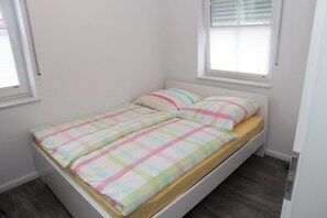 A small, single-bed room with a plain white wall, a window with blinds, and a wooden floor.
