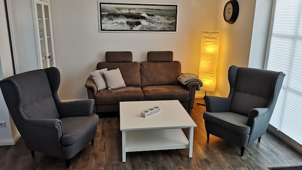 A living room with a brown sofa, two armchairs, a white coffee table, a framed picture on the wall, a lamp, and a clock. - Anchorage - Family-friendly semi-detached house in a quiet location (Krummhörn)