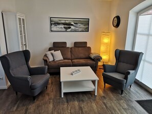 Un salón con un sofá marrón, dos sillones, una mesa de centro blanca, un cuadro enmarcado en la pared, una lámpara y un reloj.