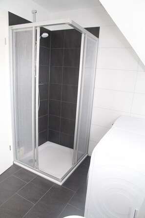Ducha en esquina con mampara de vidrio, paredes de azulejos negros y una bañera blanca.
