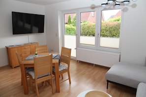 A room with a wooden dining table, chairs, a TV mounted on the wall, a radiator, and a window with a view of greenery and buildings outside. - Biene - Cozy, newly renovated vacation apartment for 3 people (Krummhörn)