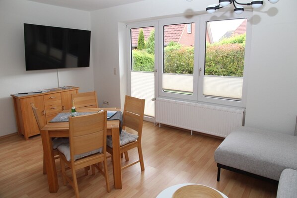Une pièce comprenant une table à manger en bois, des chaises, un téléviseur fixé au mur, un radiateur et une fenêtre donnant sur un paysage verdoyant et des bâtiments.