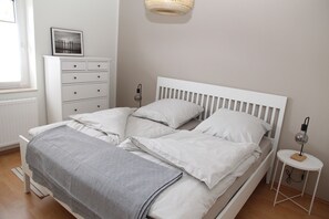 A neatly made bed with white bedding, a bedside table with a lamp, and a dresser with drawers. - Biene - Cozy, newly renovated vacation apartment for 3 people (Krummhörn)