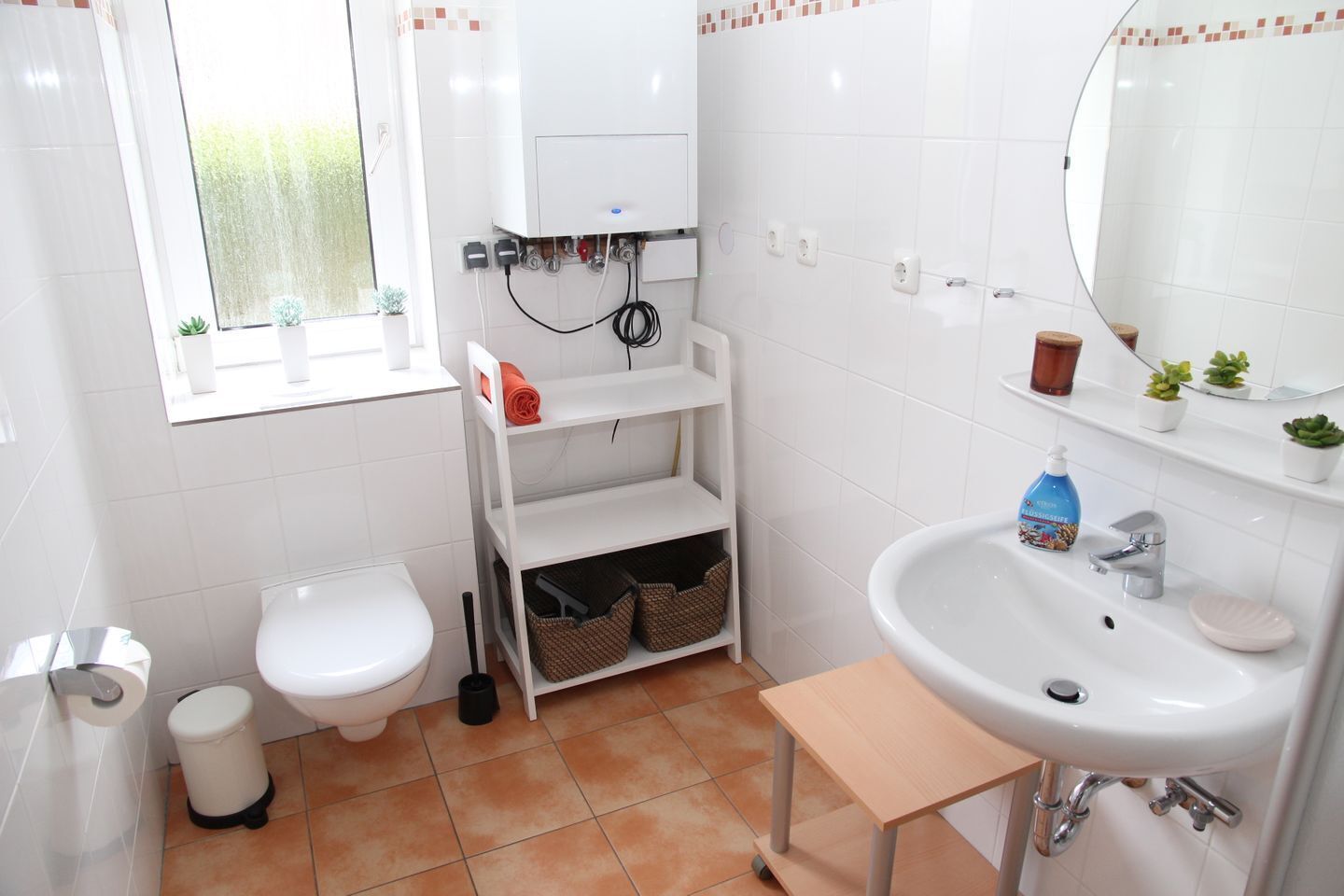 Ein Badezimmer mit Toilette, Bidet, Waschbecken und Fenster. Es gibt Regale mit Körben und einen kleinen Tisch.