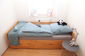 Une petite pièce chaleureuse, avec un lit en bois, une petite table sur laquelle se trouve une lampe, et une fenêtre ornée de deux tableaux encadrés.