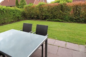 Une terrasse dotée d’une table à plateau en verre et de deux fauteuils en osier, entourée d’une pelouse bien entretenue et de haies.