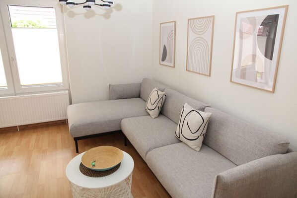 A modern living room with a grey sectional sofa, two decorative pillows, a round coffee table, and three framed artworks on the wall. - Biene - Comfortable, recently renovated vacation apartment for 3 people (Krummhörn)