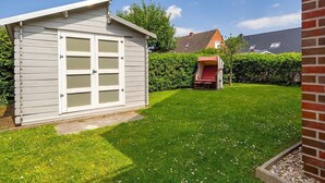 A small, single-story shed with a white exterior and a garage door, surrounded by a well-maintained lawn and a red chair. - Gode Stuuv - Our "good room" (75sqm) for 4 persons centrally located in Greetsiel (Krummhörn)