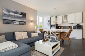 A modern living room with a grey sofa, white coffee table, and a dining area with wicker chairs. - Gode Stuuv - Our "good room" (75sqm) for 4 persons centrally located in Greetsiel (Krummhörn)