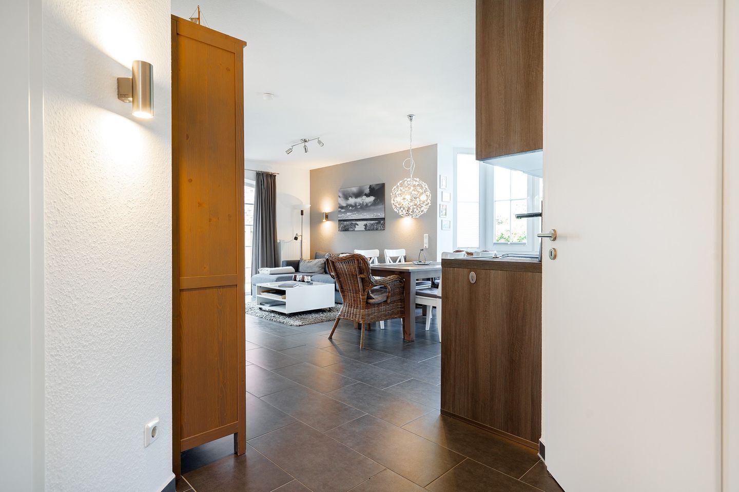 Una cocina moderna con baldosas oscuras, armarios de madera y un comedor con mesa blanca y sillas de mimbre.