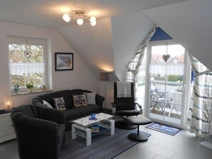 Un salon moderne avec un canapé noir, une table basse et un fauteuil noir. Une grande fenêtre offre une vue sur l’extérieur.
