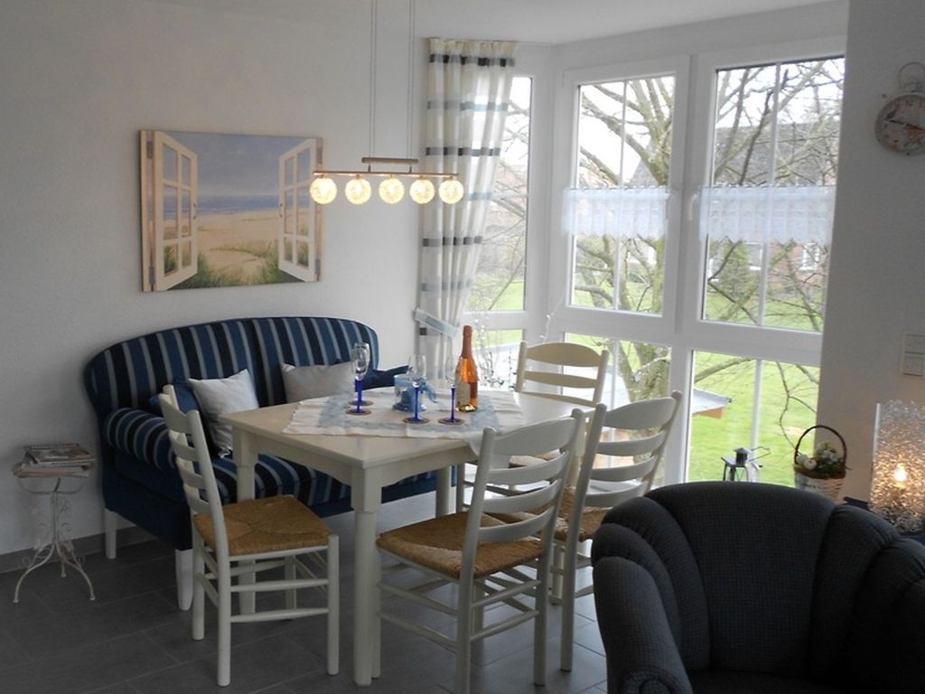 A dining area with a white table, striped blue and white sofa, and white chairs. There is a painting of open doors on the wall, and a window with a view of trees.