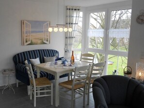 Ein Essbereich mit einem weißen Tisch, einer blau-weißen gestreiften Couch und weißen Stühlen. An der Wand hängt ein Gemälde von offenen Türen, und ein Fenster bietet Blick auf Bäume.