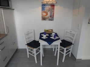 A small dining area with a table set for two, white chairs, a blue tablecloth, and a wall-mounted lamp.