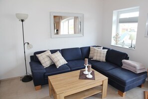 Un salon avec un canapé bleu foncé, deux coussins à carreaux, une table basse en bois, un lampadaire, un miroir et une fenêtre donnant sur l’extérieur.