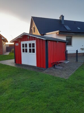 Un abri rouge et blanc avec une porte blanche, situé dans une zone herbeuse, avec un chemin pavé menant à une maison dont le toit est de couleur sombre.