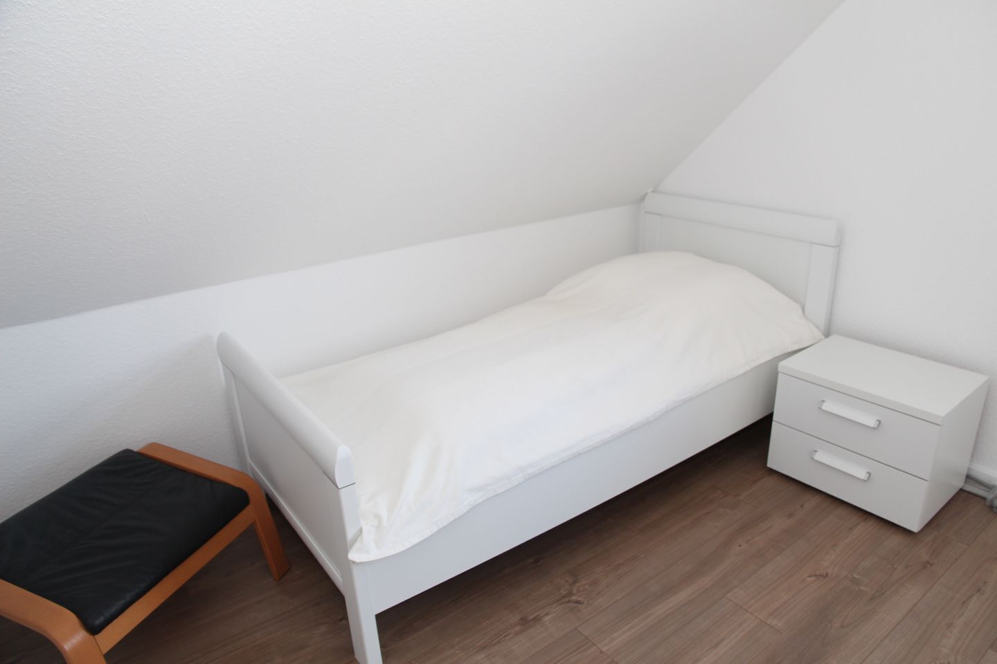 A small, white, single-bed room with a wooden bench and a nightstand with drawers.
