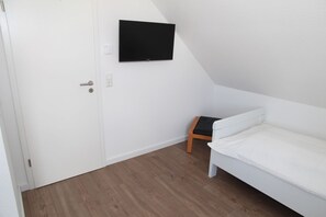 Une petite pièce blanche, avec un lit simple, un téléviseur fixé au mur et une porte dotée d’une poignée.