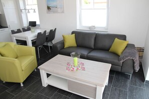 A modern living room with a grey sofa, yellow cushions, a yellow armchair, a white coffee table, and a dining area with a white table and chairs. - Matjes - Apartment (55sqm) for 4 persons and dog with terrace in Greetsiel (Krummhörn)