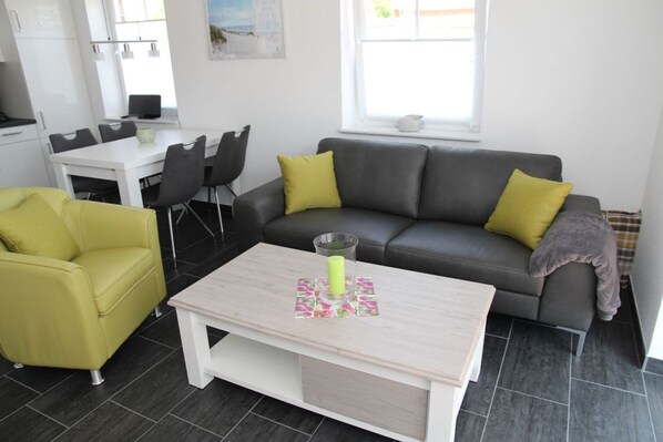 Una sala de estar moderna con un sofá gris, cojines amarillos, un sillón amarillo, una mesa de centro blanca y una zona de comedor con una mesa y sillas blancas.