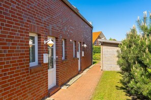 Un bâtiment en briques rouges, avec une petite structure en bois attenante, un chemin pavé et une pelouse bien entretenue agrémentée de pins.