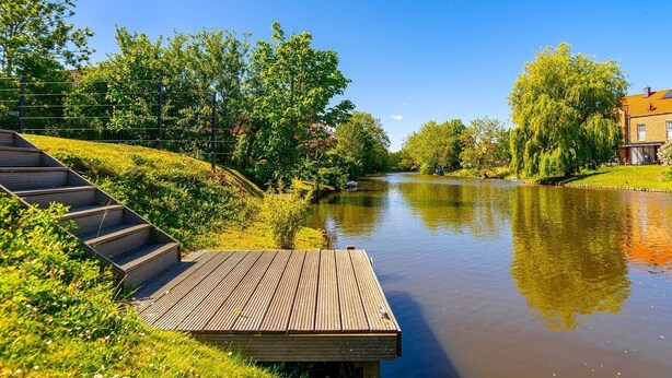 A wooden deck by a calm river with a bridge in the background and a building with a red roof. - Leo - Schöne Erdgeschosswohnung mit Hund in Wassernähe (Krummhörn)