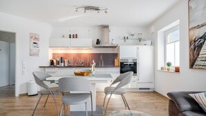 A modern kitchen with white cabinets, a central island, and a dining area with two grey chairs. - Leo - Schöne Erdgeschosswohnung mit Hund in Wassernähe (Krummhörn)