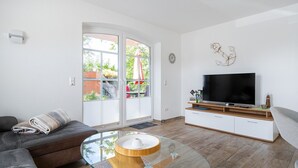 A modern living room with a glass coffee table, leather sofa, and a flat-screen TV on a wooden TV stand. - Leo - Schöne Erdgeschosswohnung mit Hund in Wassernähe (Krummhörn)