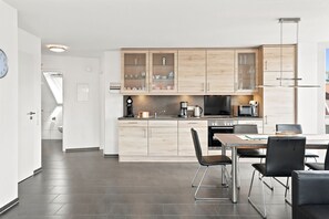 A modern kitchen with wooden cabinets, a dining area with a table and chairs, and a visible clock on the wall. - Meerzeit - Schöne Obergeschosswohnung mit Wasserzugang (Krummhörn)