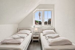 A small, single-room hotel with two beds, a window with a view of buildings, and a simple bedside table with a lamp. - Meerzeit - Schöne Obergeschosswohnung mit Wasserzugang (Krummhörn)