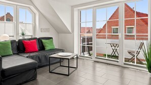 Un salon moderne avec un canapé noir, des coussins verts et rouges, une petite table et de grandes fenêtres donnant sur des bâtiments en briques rouges.