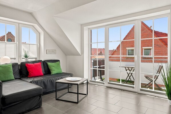 A modern living room with a black sofa, green and red cushions, a small table, and large windows offering a view of red brick buildings. - Meerzeit - Schöne Obergeschosswohnung mit Wasserzugang (Krummhörn)