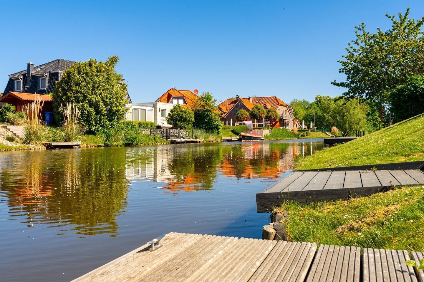 Ein ruhiger Fluss mit Häusern am gegenüberliegenden Ufer, einer hölzernen Anlegestelle und blauem, wolkenlosem Himmel.