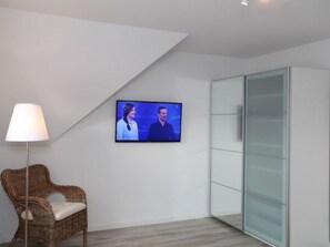 Una habitación moderna con televisión de pantalla plana montada en la pared, una silla de mimbre con cojín y un aparador de vidrio.