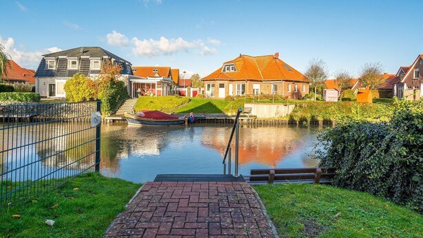 A canal with a boat docked, surrounded by houses with red roofs and a grassy area with a brick pathway.