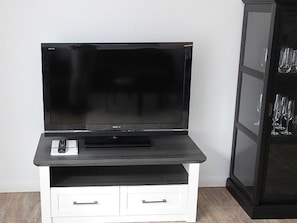 Un téléviseur à écran plat posé sur un meuble en bois foncé, avec une télécommande et un tiroir blanc en dessous.