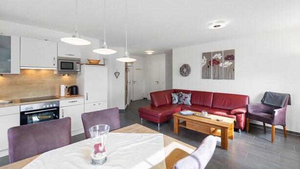 Eine moderne Küche mit weißen Schränken, einer Mikrowelle und einem Herd. Es gibt eine Essecke mit einem Tisch, Stühlen und einem roten Sofa.