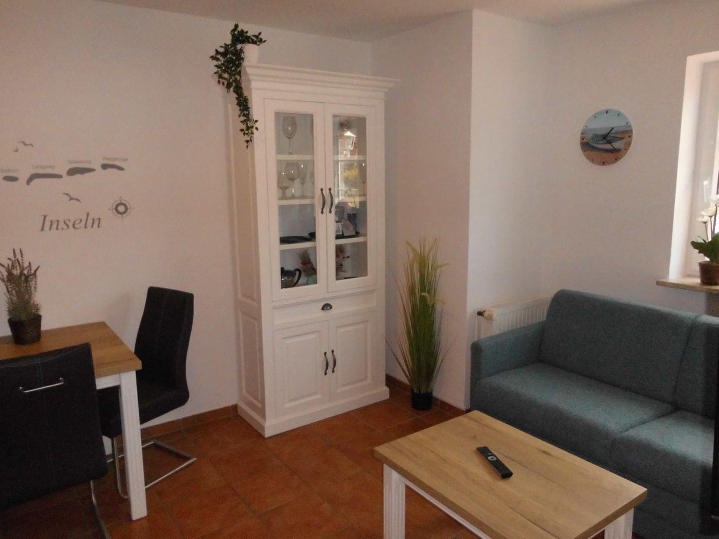 A living room with a blue sofa, a wooden coffee table, a cabinet with glass doors, and a wall with a compass and the word "Inseln".