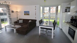 Una sala de estar moderna con un sofá marrón, una mesa de comedor con sillas y una zona de cocina con armarios y electrodomésticos blancos.