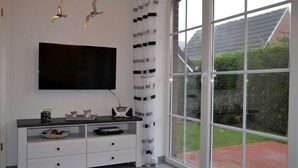 Un salón moderno con un televisor de pantalla plana, una consola blanca con objetos decorativos y un ventanal que ofrece vistas a un jardín y una casa.