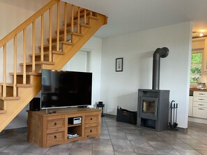 Un salon avec un escalier en bois, un téléviseur à écran plat posé sur un meuble en bois, une cheminée et un coin cuisine visible à travers une porte ouverte.