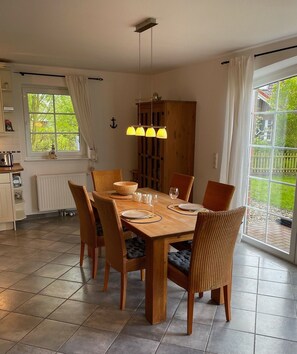 A dining area with a wooden table, chairs, a hanging light fixture, a view through a window, and a kitchen area with a radiator. - Deichbrise - Deichbrise Vacation Home: A Relaxing Family Vacation in Greetsiel with (Krummhörn)
