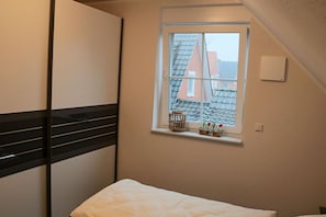 Ein Schlafzimmer mit Bett, einem Fenster mit Blick auf Gebäude, einem Regal mit Dekoration und einem Lichtschalter.