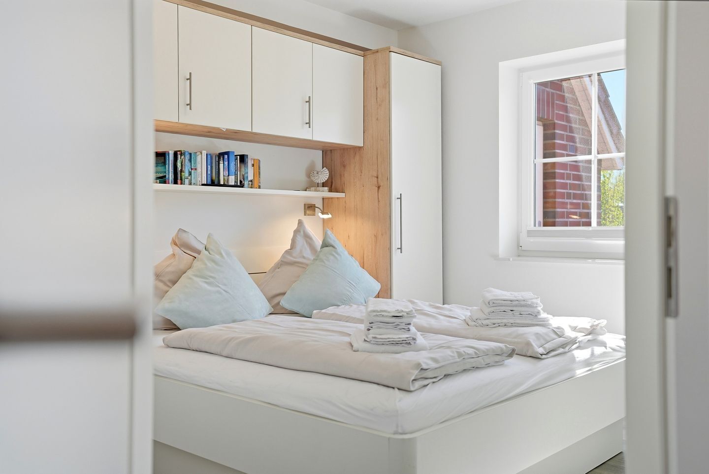 Una camera da letto con un letto, cuscini, un comodino con asciugamani, una finestra e una libreria.