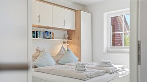Ein Schlafzimmer mit einem Bett, Kissen, einem Nachttisch mit Handtüchern, einem Fenster und einem Bücherregal.