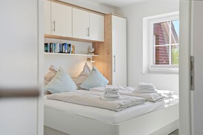 Un dormitorio con cama, almohadas, una mesita de noche con toallas, una ventana y una estantería.