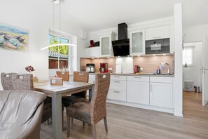 A modern kitchen with white cabinetry, a black range hood, and a dining area with wicker chairs and a table. - Nautilus - Apartment (55sqm) for 4 persons with terrace and sep. Guest WC (Krummhörn)