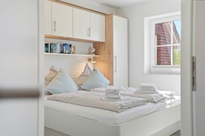 A bedroom with a bed, pillows, a bedside table with towels, a window, and a bookshelf.
