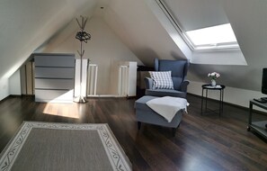 Une chambre de style loft moderne, avec un plafond en pente, une grande fenêtre, un fauteuil gris, une petite table avec un vase et une lampe sur pied.
