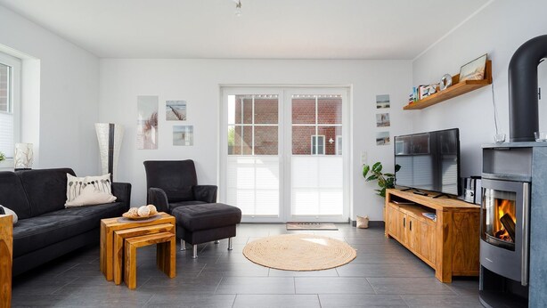 A modern living room with a black sofa, a wooden coffee table, a black armchair, a TV, a fireplace, and a large window with a view of a brick building. - Löwenzahn - Apartment (70sqm) for 4 persons with terrace and sep. Guest WC (Krummhörn)